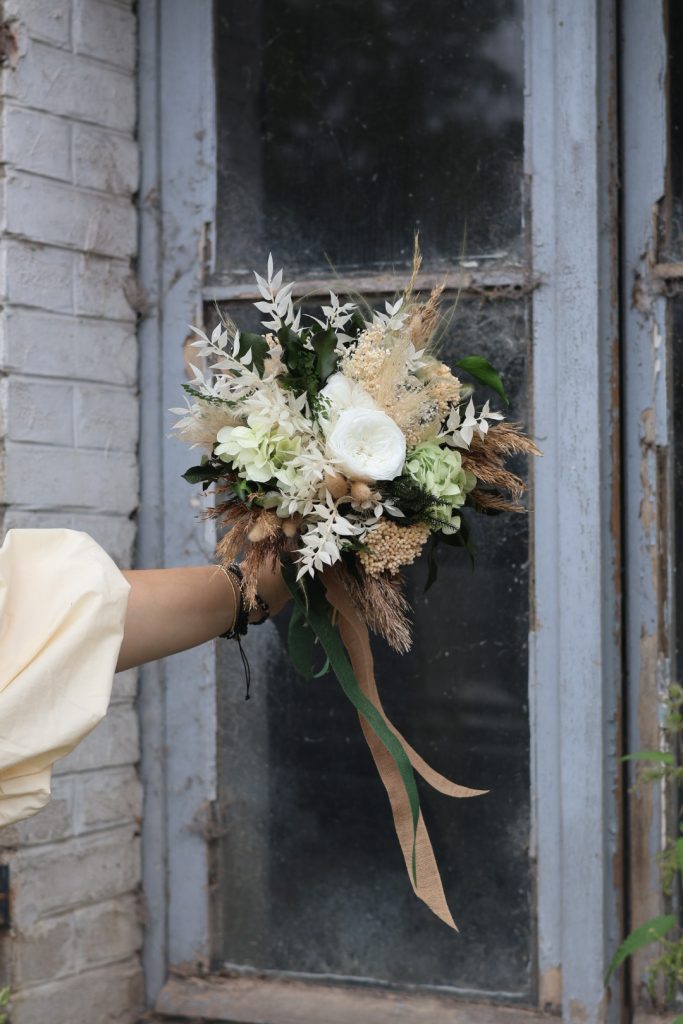 Un joli bouquet de fleurs dans les tons beiges et blancs tenu par la main de la fleuriste devant une fenêtre