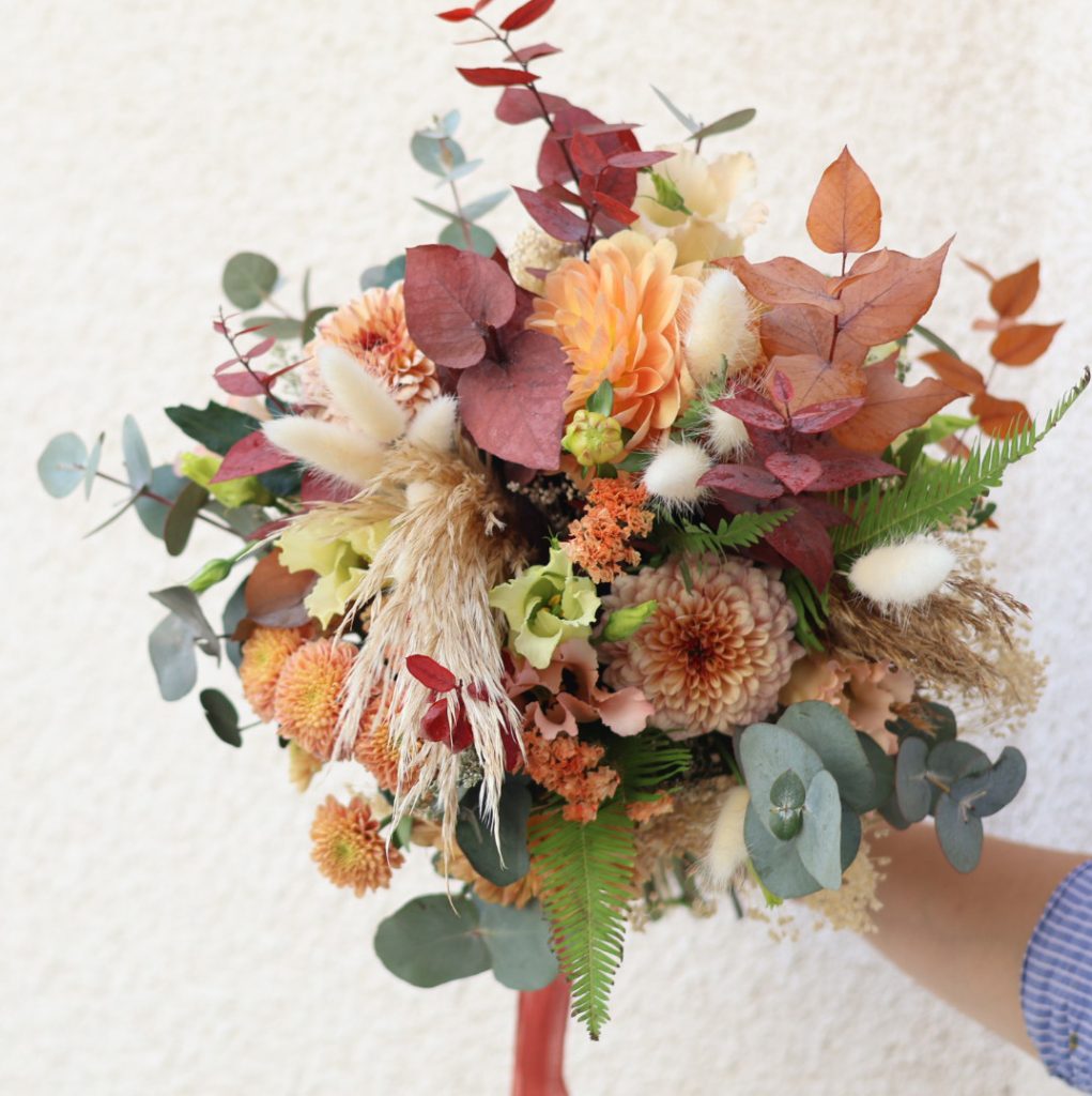 Un bouquet de fleurs aux couleurs d'automne avec des fleurs fraîches et fleurs séchées, tenu devant un mur par la main de la fleuriste