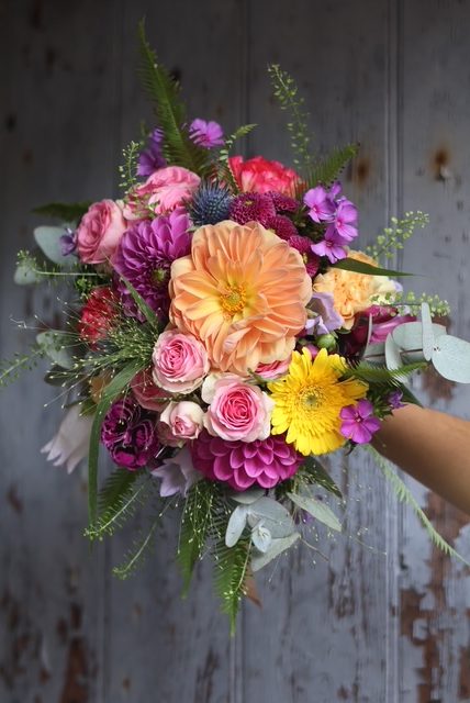 Un magnifique bouquet de fleurs coloré : jaune, orange, rose et violet, tenu par la fleuriste devant un fond sombre