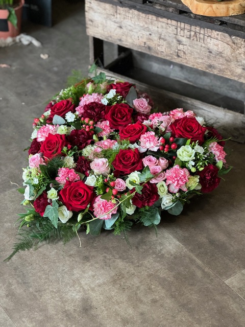 Un coussin funéraire en forme de coeur, composé de roses rouges et roses ainsi que des oeillets et autres fleurs