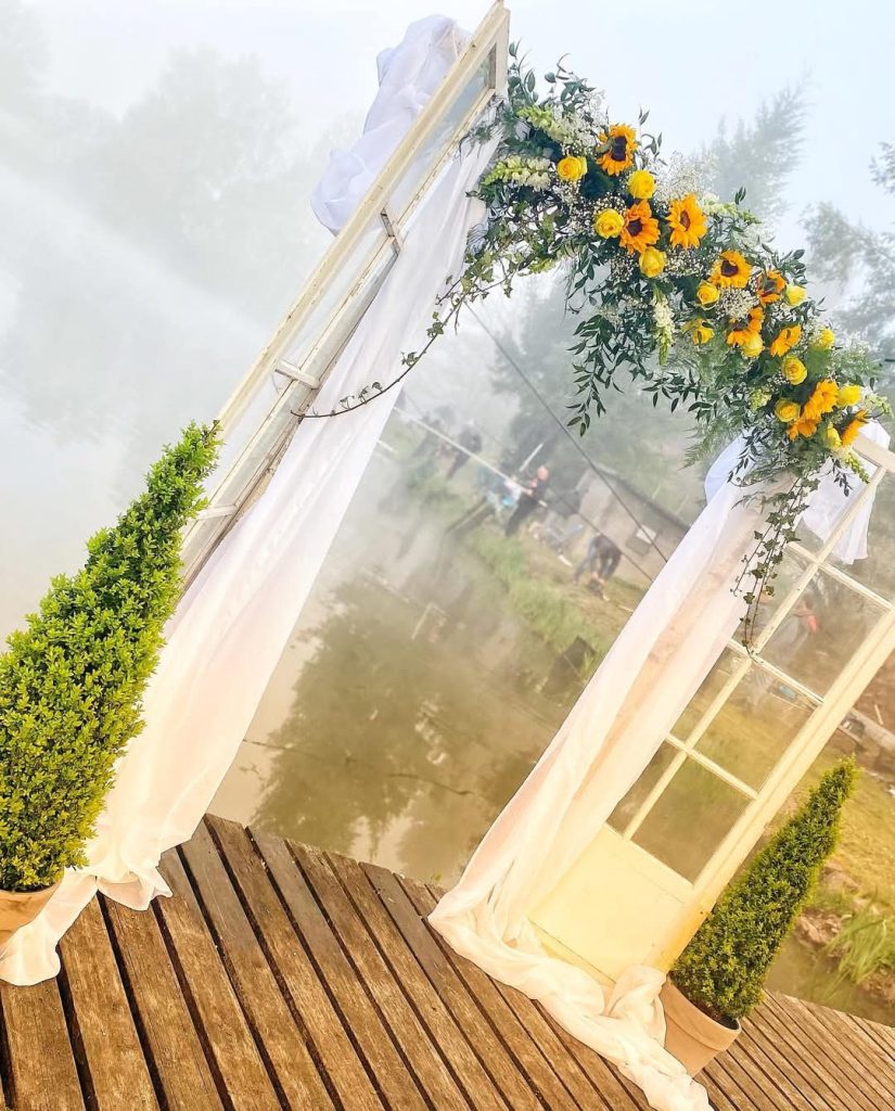Création sur mesure pour un mariage dans les Vosges : Une arche fleurie composée de deux portes vitrées sur les extrémités, recouverte de voiles blanc, feuillage et des fleurs jaunes, notamment des tournesols.