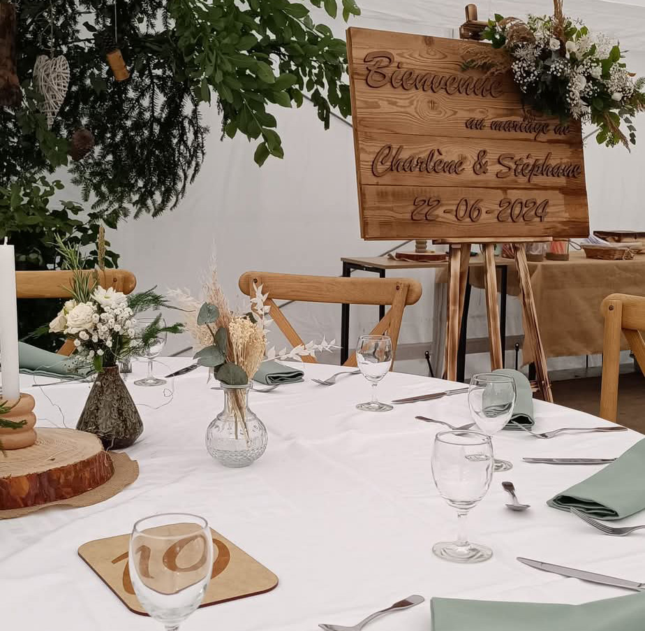 Une table décorée de vase garnis de fleurs et centre de table en bois, avec un panneau de bienvenue au mariage fleurit en arrière plan décoré par les fleuristes.