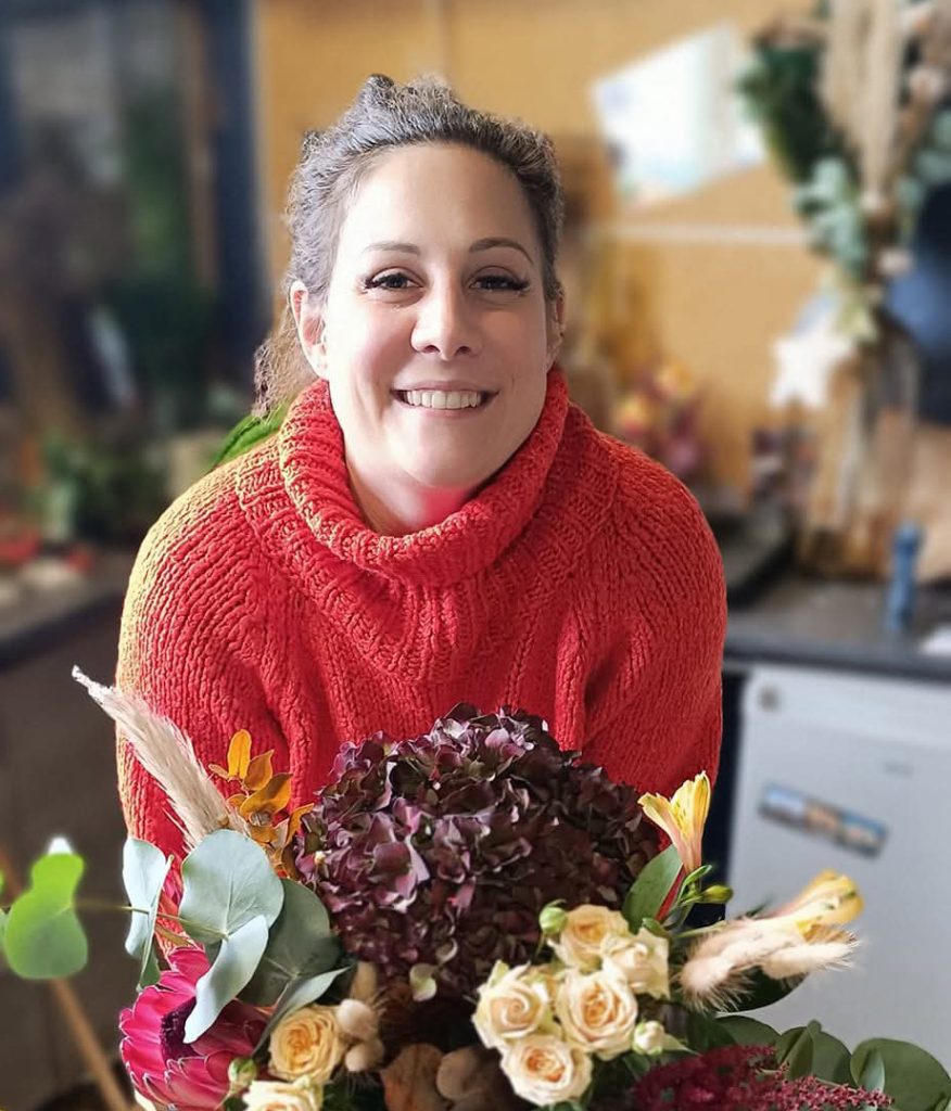 Photo de la fleuriste Pauline qui tient un joli bouquet de fleurs et qui fait un grand sourire dans la boutique Un Brin d'Folie à Arches