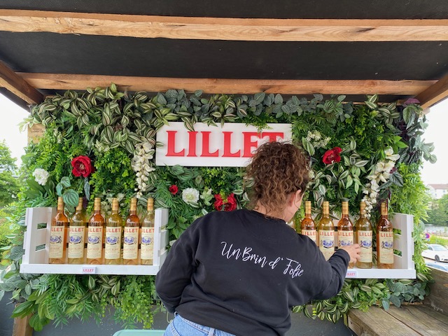 La fleuriste Pauline est de dos, elle porte un pull floqué "Un Brin d'Folie", et est en train de composer une décoration végétale sur un stand pour une entreprise de boisson