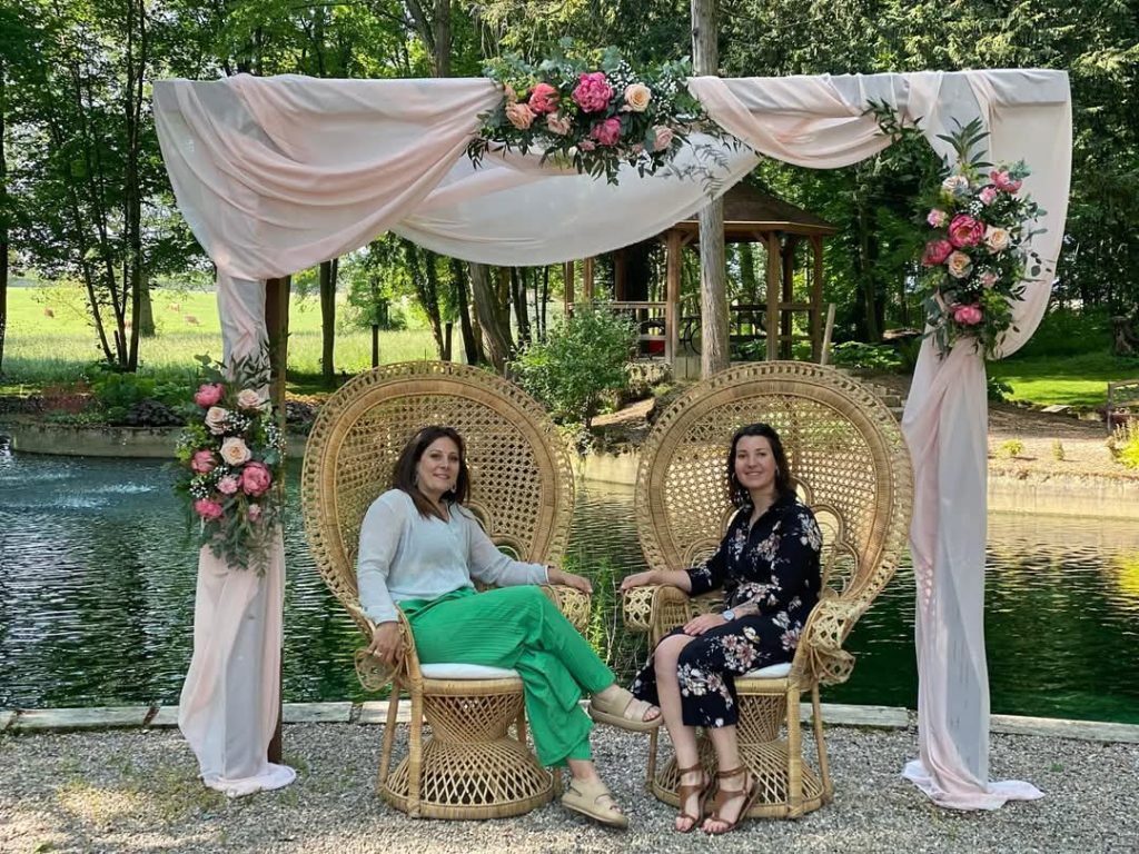 Les deux fleuristes sont assises sur de belles chaises en rottin sous une arches décorée de fleurs et de voiles roses avec une rivière en arrière plan lors d'un mariage dans les Vosges