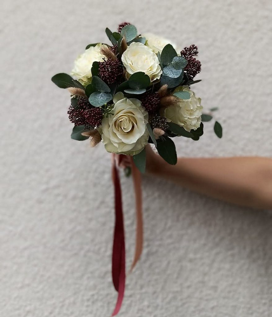 Un petit bouquet de mariée rond blanc et vert attaché par un ruban et tenu par la fleuriste devant un mur