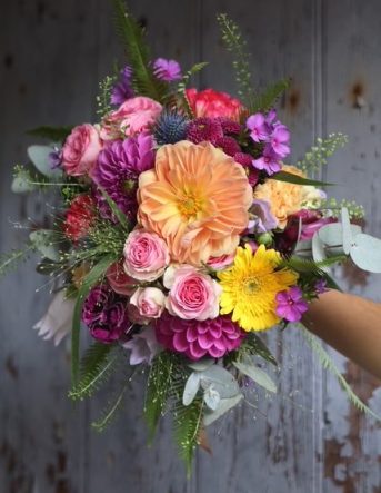 Un magnifique bouquet de fleurs coloré : jaune, orange, rose et violet, tenu par la fleuriste devant un fond sombre