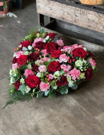 Un coussin funéraire en forme de coeur, composé de roses rouges et roses ainsi que des oeillets et autres fleurs