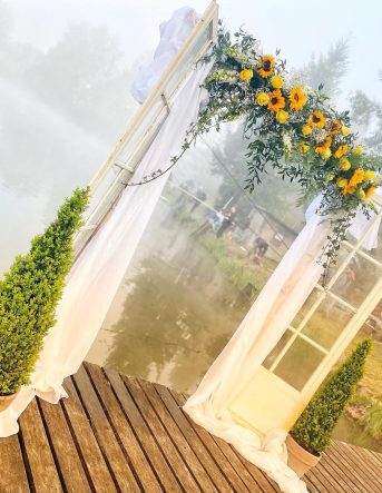 Création sur mesure pour un mariage dans les Vosges : Une arche fleurie composée de deux portes vitrées sur les extrémités, recouverte de voiles blanc, feuillage et des fleurs jaunes, notamment des tournesols.