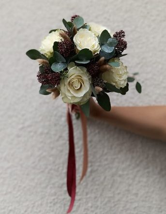 Un petit bouquet de mariée rond blanc et vert attaché par un ruban et tenu par la fleuriste devant un mur